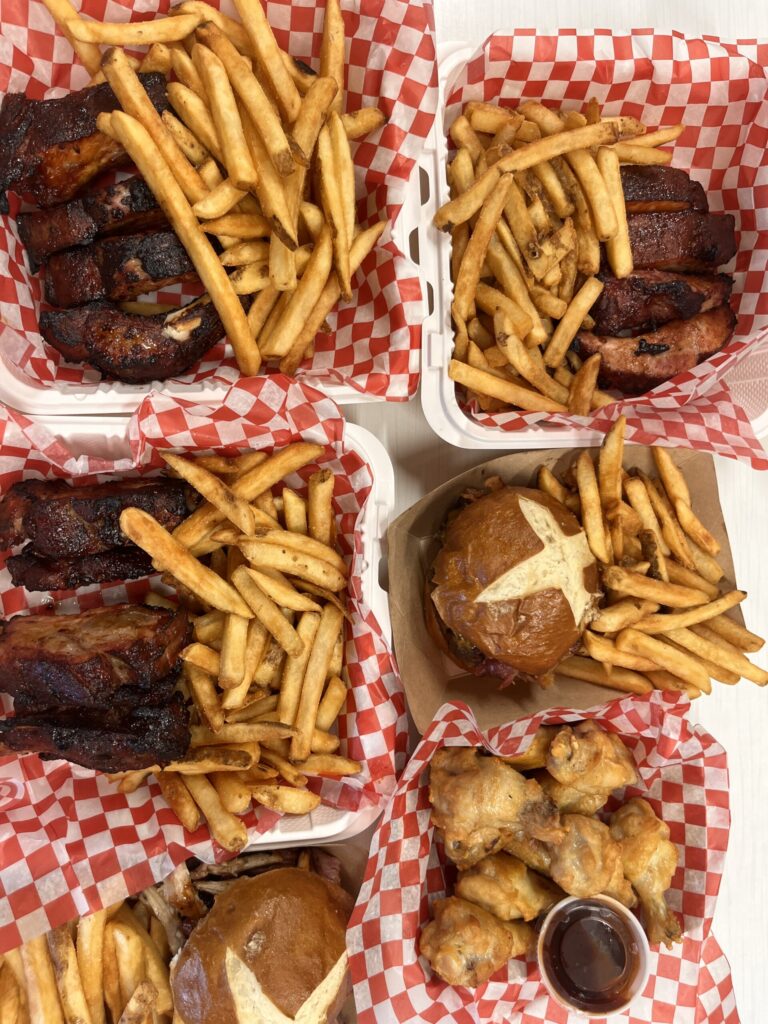 Six takeout orders set next to one another - three orders of ribs, two burger platters, and chicken wings. The meals are in takeout containers, with red and white liners.